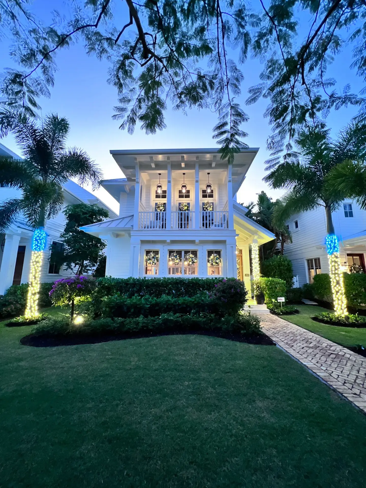 White coastal Naples home with lit palms and holiday wreaths at dusk
