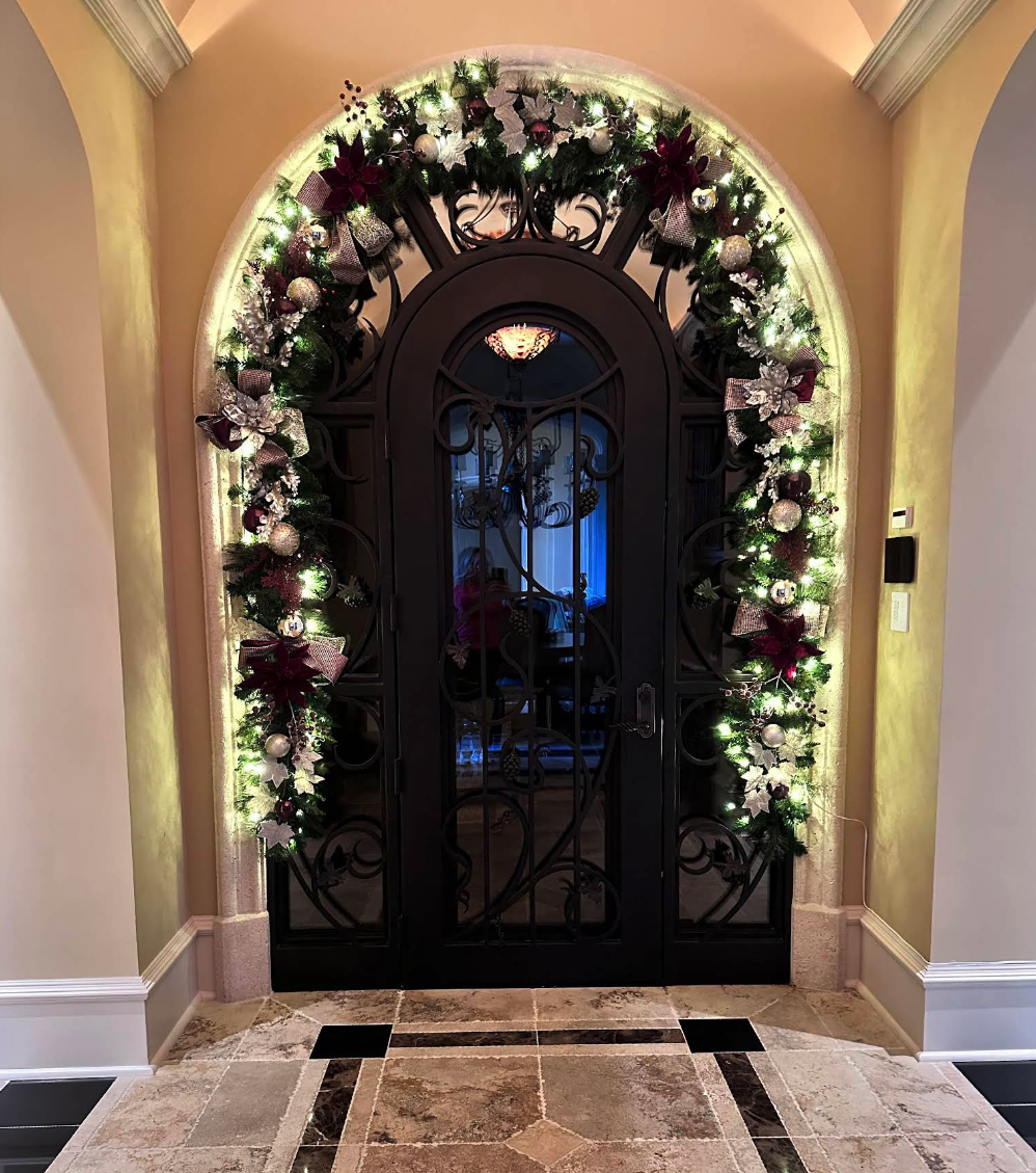 Elegant arched front door decorated with lit garland, ornaments and red bows